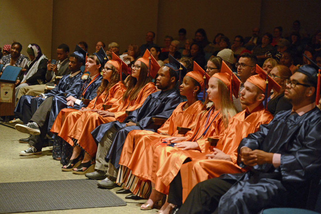 Group of 10 students in either orange or black graduation gowns, seated, with audience behind them.