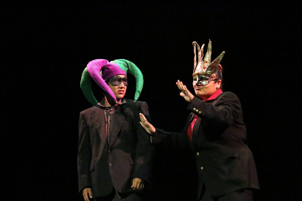 Two high school males in black suits and carnivale masks on dark stage.