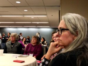 Three adults at a table watch a speaker, with many others in the background seated at tables.