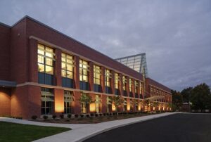 Rectangular brick building with large, lit windows in front of darkening sky.